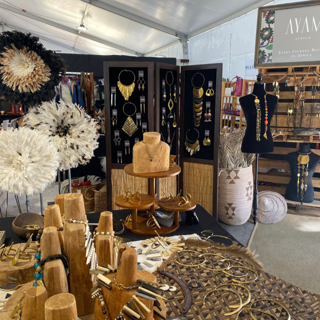 # Jewelry Display and Retail Booth Setup

This is a modern jewelry retail display booth featuring wooden jewelry stands and display fixtures in natural pine and walnut tones, circa 2020s. The setup includes multiple black velvet-backed display panels showcasing gold-toned and mixed-metal necklaces, earrings, and bracelets, complemented by decorative woven natural fiber wall hangings and sculptural fiber art pieces in cream and black. Notable features include tiered wooden ring stands, numerous wooden bust forms for necklace display, organized hanging jewelry displays on black panels, and branded signage reading "AYAN," suggesting this is a contemporary jewelry designer's pop-up or market booth with an emphasis on artisanal gold jewelry and bohemian aesthetic.