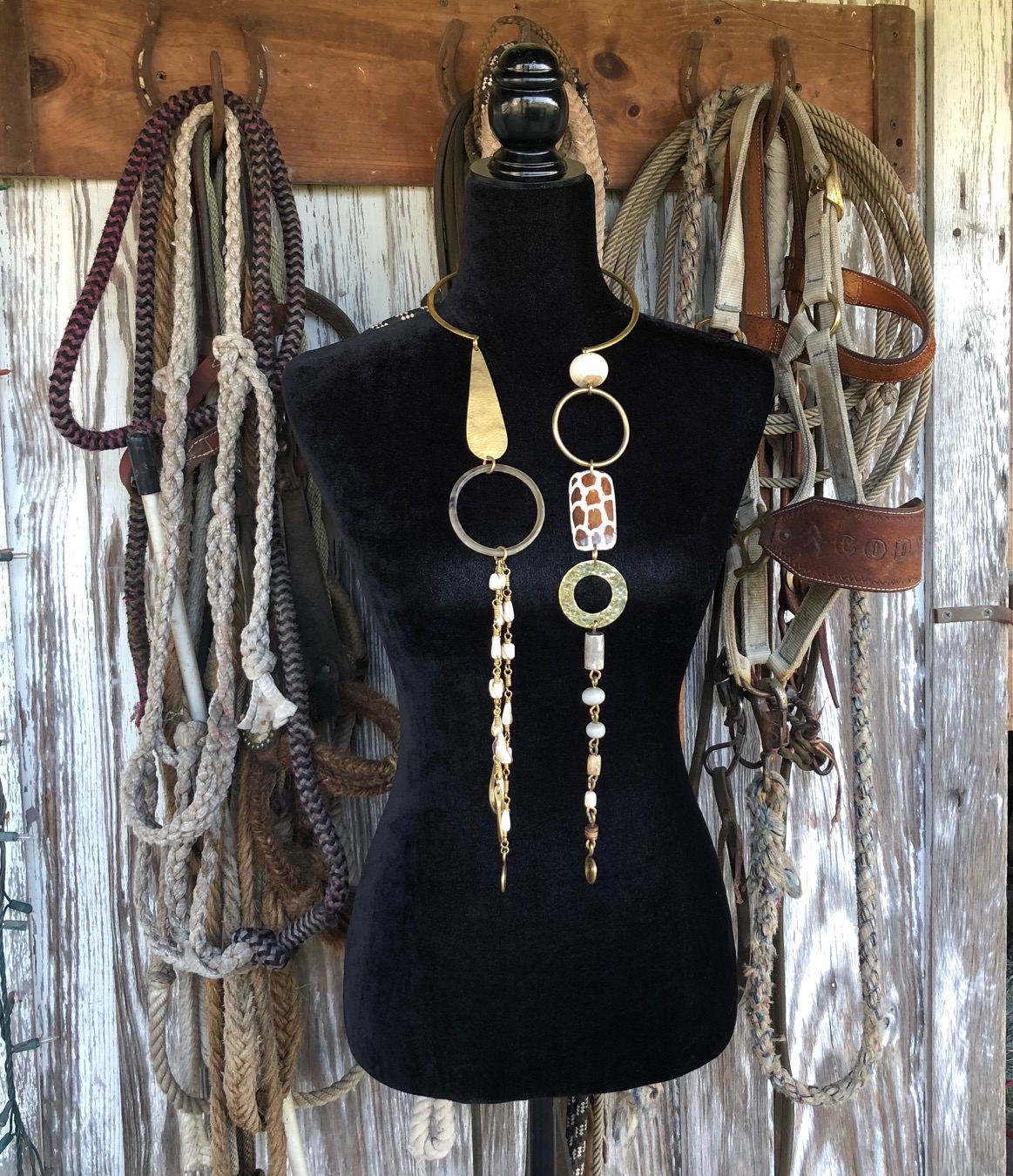 # Vintage Rope and Tack Display with Mannequin

This is a decorative display featuring a black fabric dress form mannequin mounted on a rustic wooden panel with a two-tone finish (natural wood top and whitewashed lower section). The mannequin is styled with long chain link earrings featuring brass rings, pearl accents, and geometric elements in a bohemian aesthetic, and is surrounded by multiple coiled ropes and leather horse tack in natural tan, cream, burgundy, and brown tones hung from wooden pegs. The arrangement creates a curated vintage or Western-inspired display piece, likely from the late 20th century or contemporary made to look vintage, showcasing equestrian and rustic home décor styling.